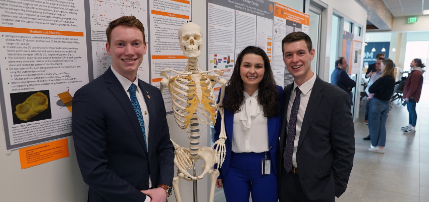 Students stand in front of research posters with anatomical skeleton.