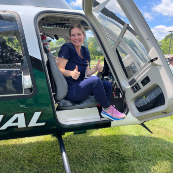 A smiling woman dressed in navy blue scrubs gives two thumbs up while sitting inside the open door of a helicopter with