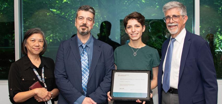 Four individuals standing together, with one holding a certificate of recognition. The group includes three men and one woman, all dressed in professional attire, posing for a photo in front of a window with greenery outside.