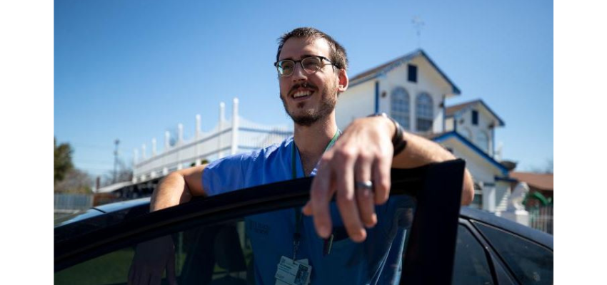 A physician wearing blue scrubs and glasses, standing outside and leaning on a car door. The individual is smiling and appears to be enjoying a sunny day. A building with a distinctive design is visible in the background.