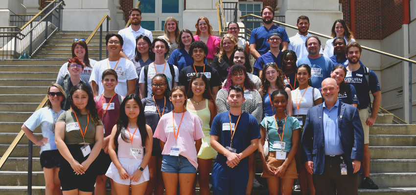 A group photo of students and faculty members standing on the steps of a building. The group includes a diverse mix of individuals, all smiling and wearing name tags.