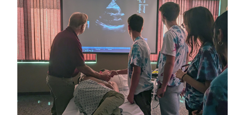 An instructor guides a students while using the ultrasound probe on a patient lying on a bed.