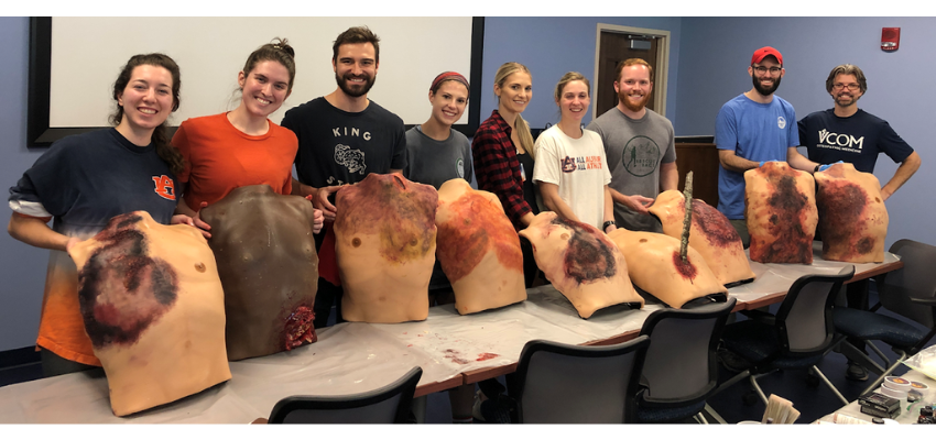 A group of students in a classroom posing with realistic, simulated injury torsos used for medical training.