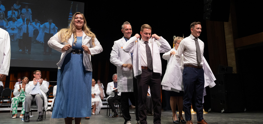 Medical students at a white coat ceremony, smiling as they put on their new white coats on stage.
