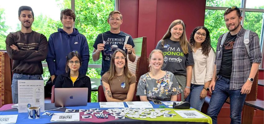 A diverse group of young adults, some standing and some sitting behind a table with informational materials, smiling and posing for the photo indoors.