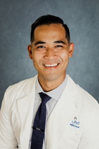 A man in a white coat and striped tie smiling in a professional headshot against a gray background.
