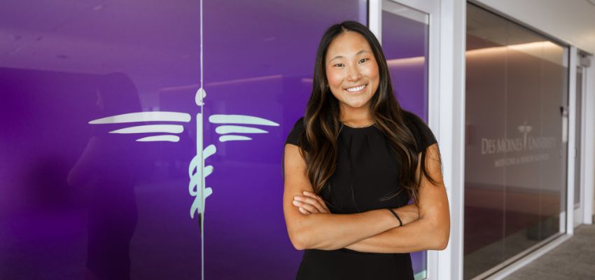 A medical student poses confidently with crossed arms in front of a university logo on a glass wall.