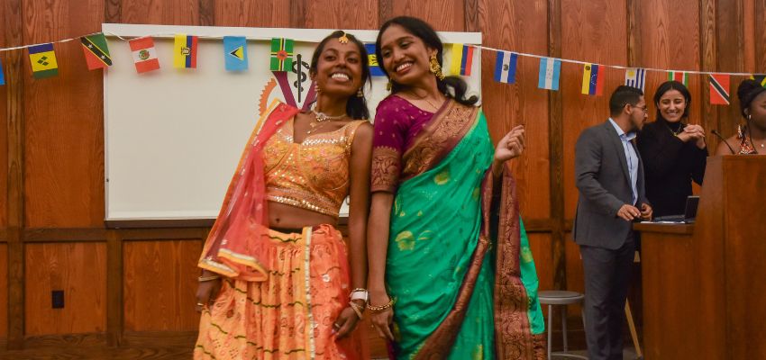 Two women in traditional cultural attire smile and pose together in front of a wall adorned with international flags.
