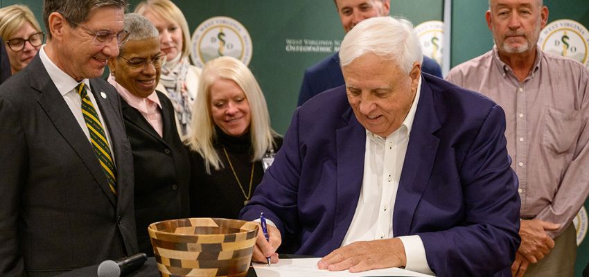 A public official signs a document at a ceremony surrounded by colleagues and officials.