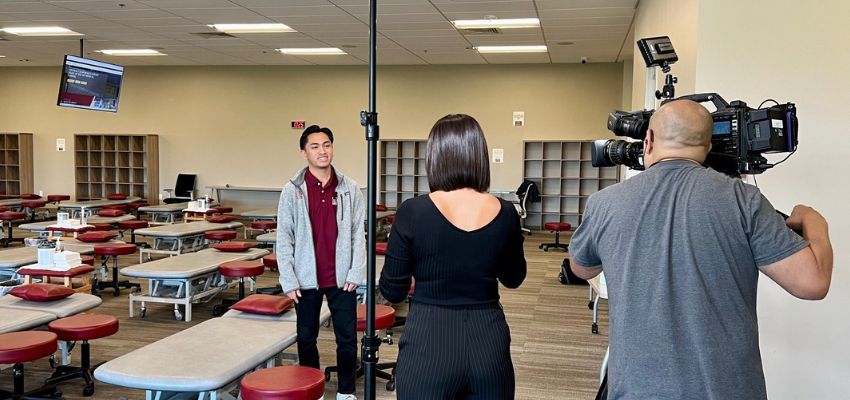 A student is interviewed on camera in a classroom filled with medical examination tables.