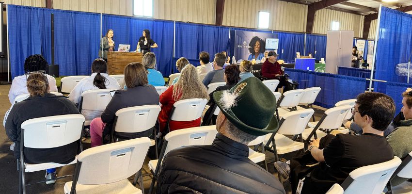A group of attendees listens to a speaker at a conference session with a blue backdrop.