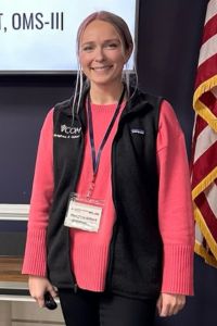 A medical student wearing a VCOM Virginia vest stands and smiles at the camera.