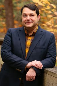 A professional headshot of a man wearing a dark blazer and standing outdoors against a fall background.