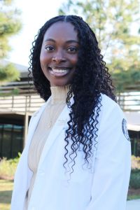 A smiling medical student in a white coat standing outdoors with a campus building in the background, signifying academic or professional pride.