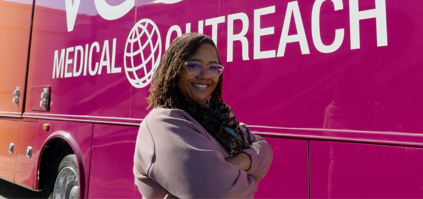 A smiling individual standing in front of a large medical outreach bus, symbolizing community healthcare efforts.