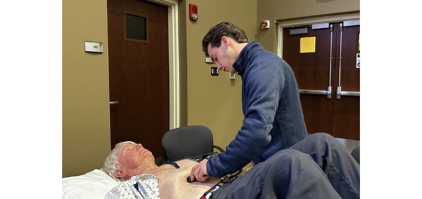 A medical student uses a handheld device to examine a patient lying on a hospital bed during a clinical skills session.