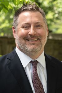 A man smiles in a professional headshot wearing a suit jacket and tie with an outdoor background.