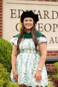 A graduate wearing academic regalia smiles while standing outdoors near a campus sign.