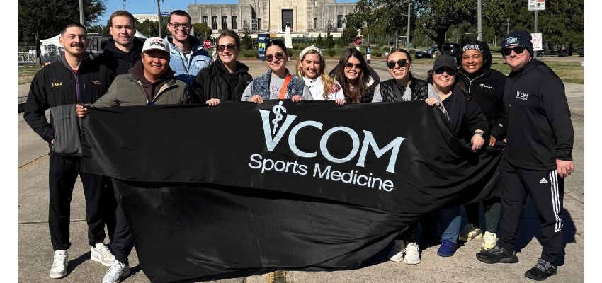A group of students and faculty pose outdoors holding a banner that reads “VCOM Sports Medicine.”