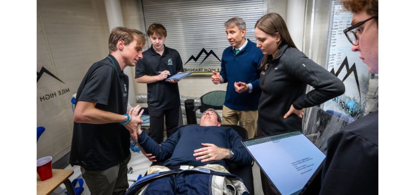 Students and faculty observe a hands-on medical training demonstration with a patient lying on an exam table.