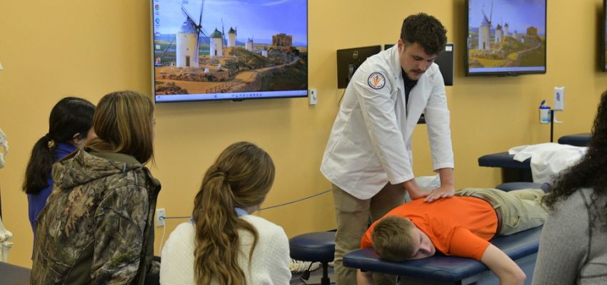 A medical student in a white coat demonstrates osteopathic manipulative treatment on a patient as classmates observe.