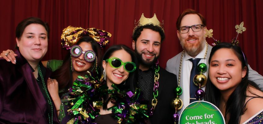 A group of people pose together in festive attire and Mardi Gras-themed accessories in front of a photo booth backdrop.