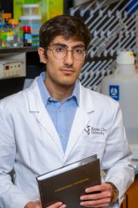 A medical student in a white coat stands in a laboratory holding a folder and looking toward the camera.