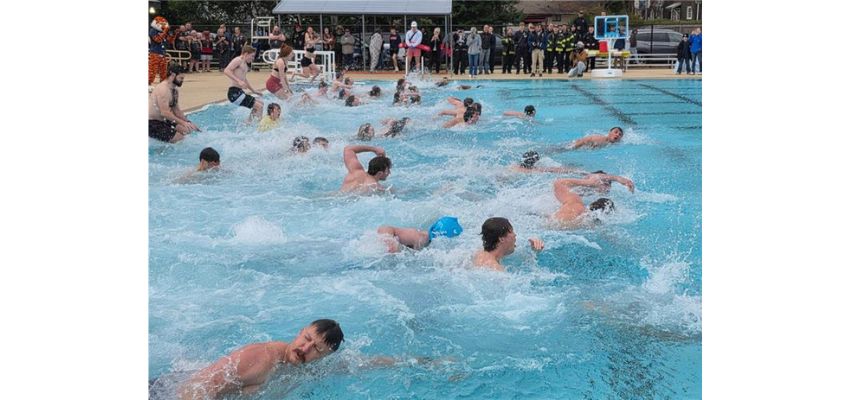 Participants swim in an outdoor pool during a large group event with spectators watching nearby.