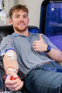A student smiles and gives a thumbs-up while donating blood in a clinical setting.