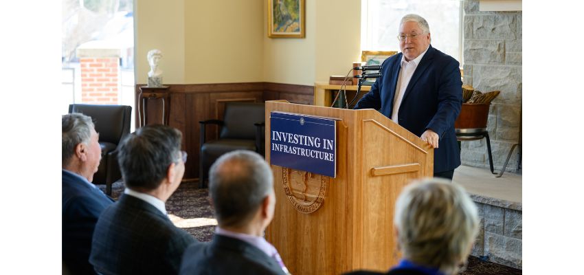 A speaker stands at a podium delivering remarks during an indoor event focused on infrastructure investment.