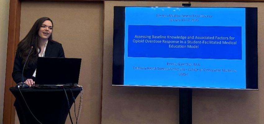 A presenter stands at a podium beside a screen displaying a research presentation slide at a medical conference.