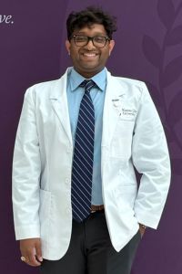 A medical student in a white coat smiles while standing in front of a purple backdrop.