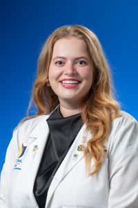 A medical student in a white coat smiles in a professional headshot against a blue background.