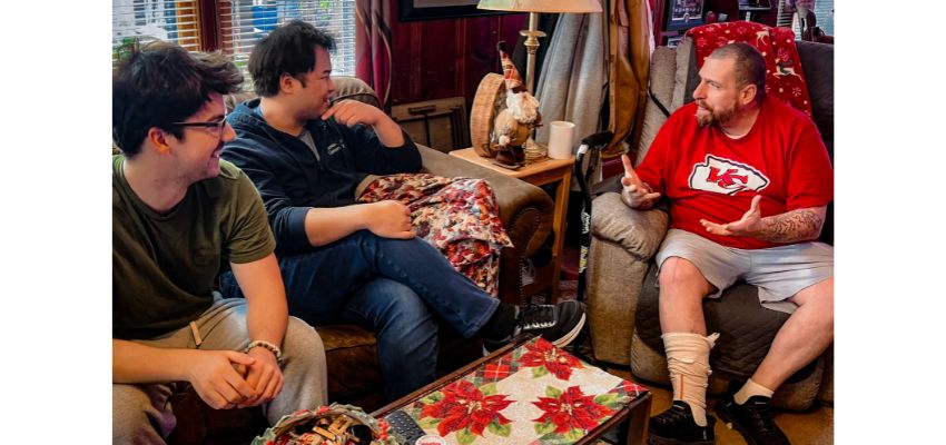 Three men sit in a living room having a conversation, with one gesturing while the others listen.