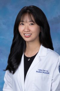 A medical student in a white coat smiles in a professional headshot against a blue studio background.