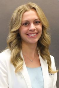 A woman in a white coat smiles in a professional headshot against a neutral background.