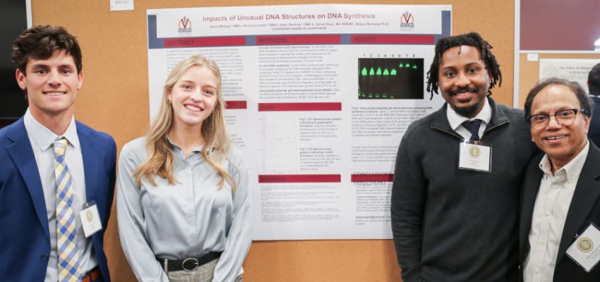 Four presenters stand smiling in front of a research poster displayed at an academic event.