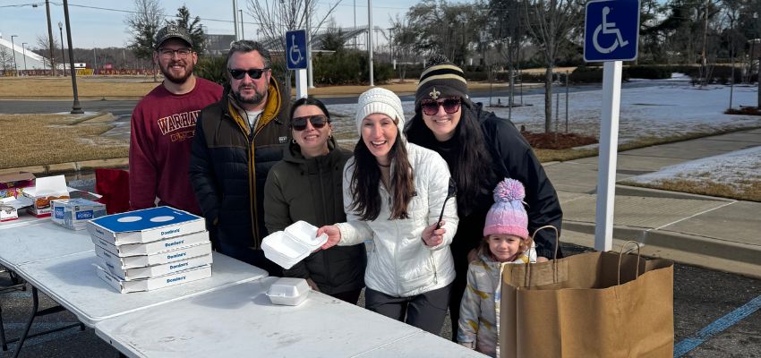 A group of people stand in a parking lot welcoming others with food.