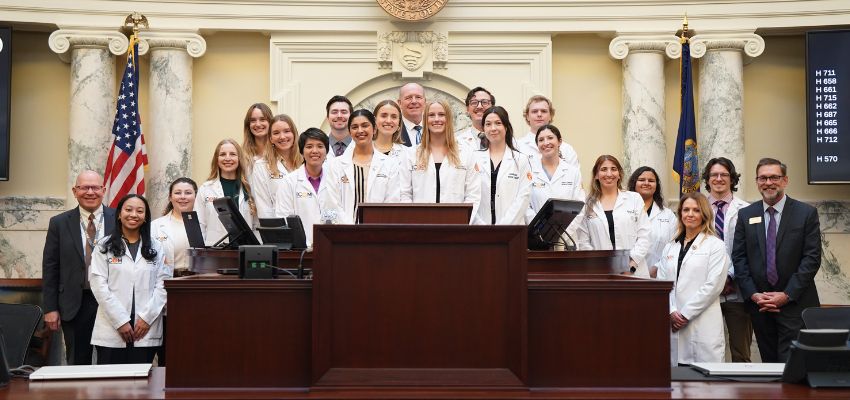 Medical students in white coats pose together inside a legislative chamber with American flags and marble columns behind them.