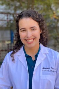 A medical student in a white coat smiles in a professional headshot taken outdoors.