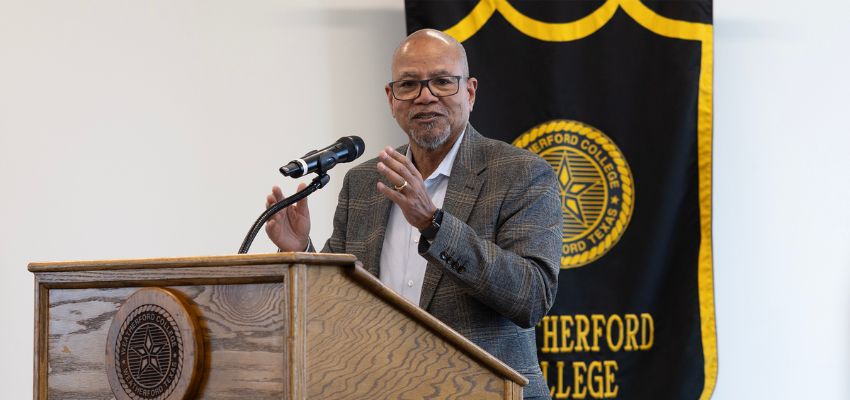 A speaker stands at a podium delivering remarks with a college banner displayed behind him.