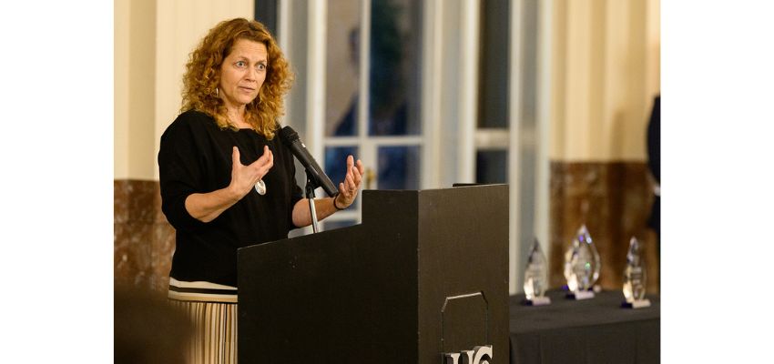 A woman speaks at a podium during a formal event with awards displayed on a table nearby.