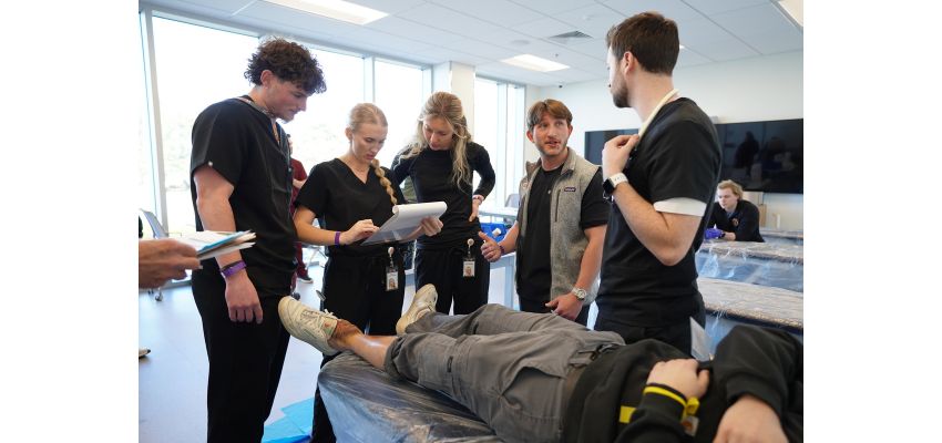 Students gather around a patient on an exam table while discussing notes during a hands-on clinical training session.