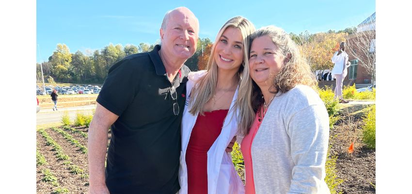 Three people stand outdoors smiling together, with one wearing a white coat, in a landscaped campus setting.