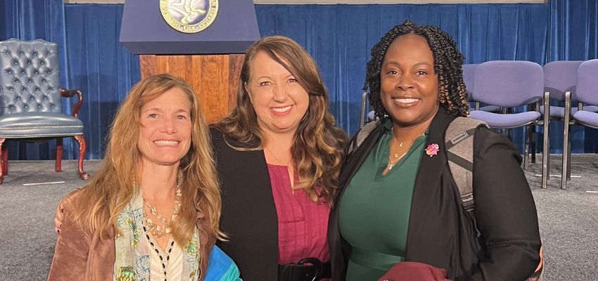 Three women stand together smiling in front of a stage with a podium and seating area.