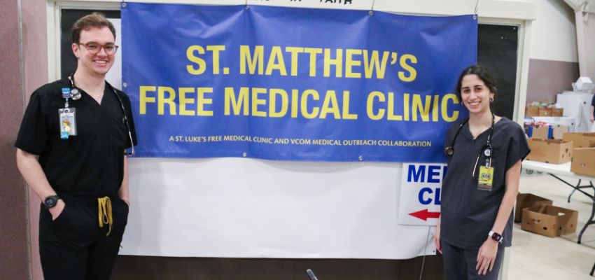 Two healthcare students stand beside a banner for St. Matthew’s Free Medical Clinic inside a clinic space.