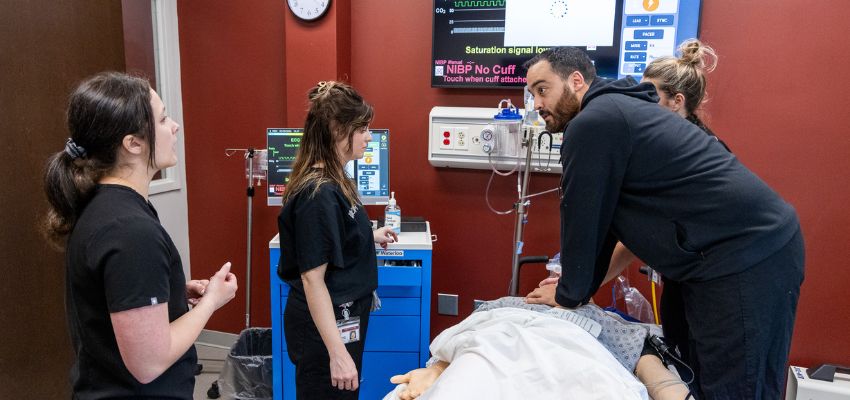 Students participate in a medical simulation as one performs a procedure on a patient while others observe.