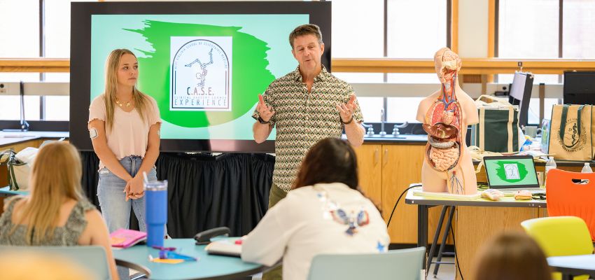 An instructor speaks to a classroom while standing near an anatomy model and presentation screen during a lesson.