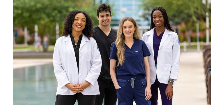 Four medical students in scrubs and white coats stand smiling together outdoors on a campus walkway.