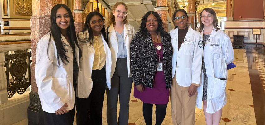 A group of medical students and a faculty member in white coats pose together inside a historic building.